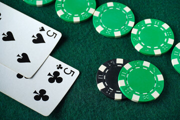 Two playing cards showing a five of clubs and a five of spades lie next to several green poker chips on a green felt table backdrop, indicating an ongoing game