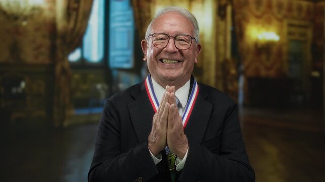 Senior man in business suit smiling with hands clasped wearing medal in museum setting indoor showcasing recognition achievement happiness elegance success museum