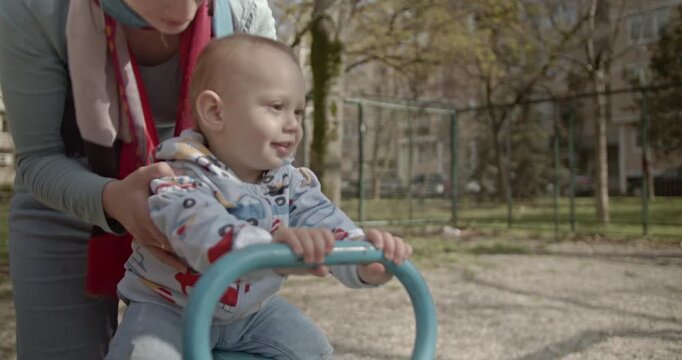 A mother gently pushes her baby boy on a seesaw in a playground. She adjusts his scarf and smiles, creating a heartwarming moment of connection.