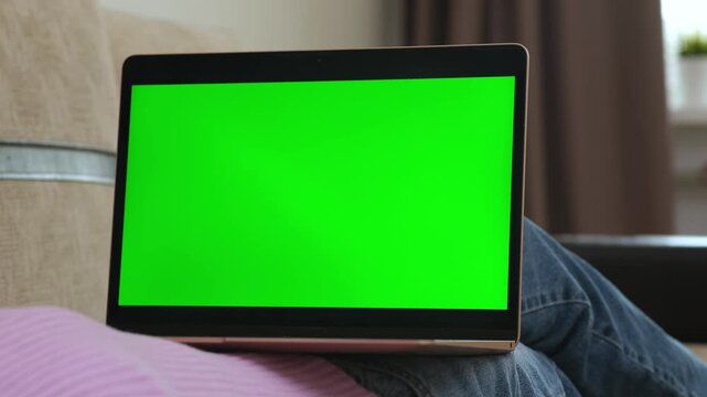 Girl watching laptop with green screen while lying on sofa at home. Female resting at couch and looking at chroma key screen