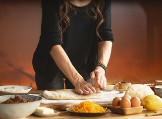 A woman presses down on pizza dough to flatten it on a counter. Around her are bowls of toppings like meat and cheese, along with eggs, dough, and other Italian food ingredients