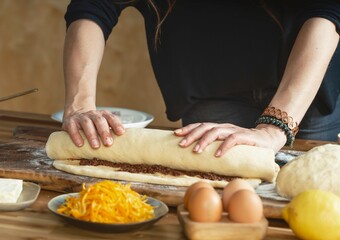 A person is preparing babka. They are rolling cinnamon filled dough on a wooden table with flour. Eggs, lemon, and shredded squash are also on the table