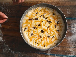 A freshly prepared cake dessert in a metal tin awaits consumption. Orange zest and dark chocolate are scattered throughout the creamy white frosting on a wooden table