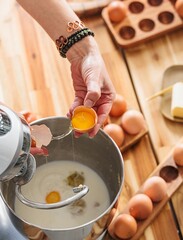 A woman prepares a baking recipe, adding egg yolks to a mixer bowl already containing ingredients. Eggs and butter are staged on the wooden counter, ready for baking