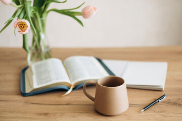 Open Bible on a wooden table and ceramic coffee mug and a vase with tulips.