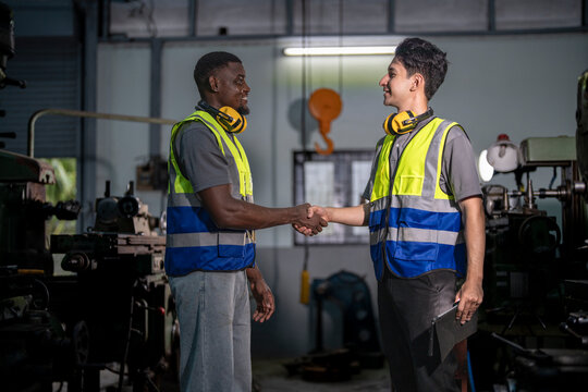 Two industrial engineers in high-visibility vests collaborate in a workshop. One holds tablet, pointing at equipment, discussing plans. Ear protection and gloves emphasize safety and professionalism.