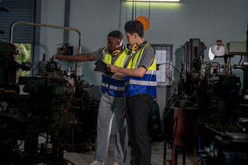 Two industrial engineers in high-visibility vests collaborate in a workshop. One holds tablet, pointing at equipment, discussing plans. Ear protection and gloves emphasize safety and professionalism.