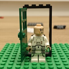 Naklejka premium A Lego minifigure in a white uniform stands at an open green door on a green baseplate, illustrating a moment of decision or cautious entry in a creative toy scenario