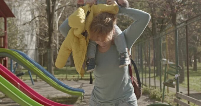 A mother gives her toddler a ride on her shoulders at the park. They are both smiling and enjoying their time together.