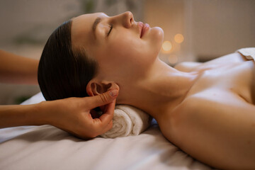 Young lady client getting ear massage by professional masseuse in spa salon, lying in relaxed atmosphere, enjoying beauty treatment. Wellness acupressure manipulations