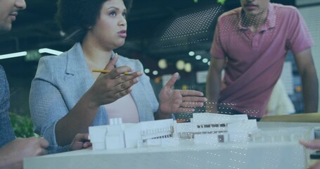 Presenting woman architect wearing blazer, gesturing with pencil at model in studio, with coworkers