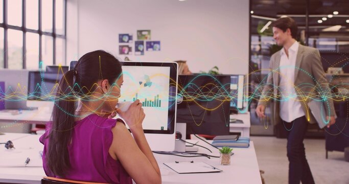 Drinking mature adult woman analyzing graphs on computer at open-plan office desk, with ceramic mug