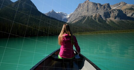 Looking woman wearing bright pink jacket sitting in canoe at turquoise mountain lake, with paddle