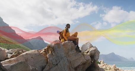 Sitting hiker in yellow jacket gazing over sea on jagged rock, with red backpack, sleeping pad