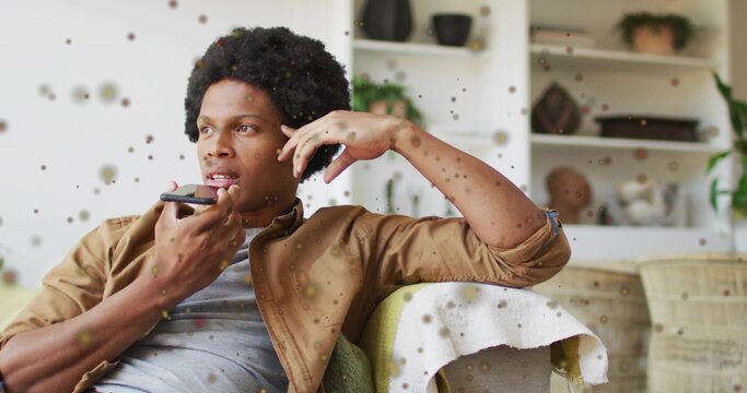 Speaking African American man touching ear, holding smartphone on sofa in living room, with shelves