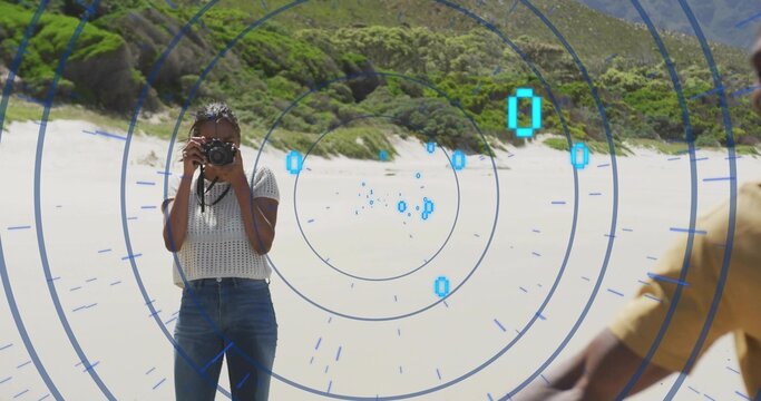 Hispanic woman pointing DSLR camera toward yellow shirt friend on beach, with digital HUD overlay - Powered by Adobe