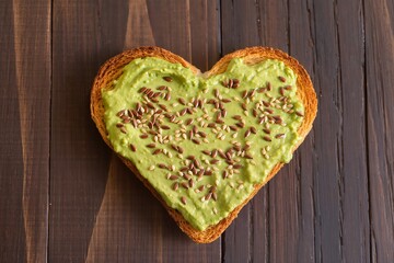 Heart-shaped toasted bread spread with avocado paste and sprinkled with flax and sesame seeds on a dark wooden background, top view