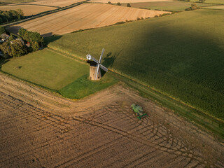 Obraz premium Aerial view of historic windmill beside harvested farmland and agricultural machinery in golden fields, rural countryside landscape with traditional architecture and seasonal farming activity 