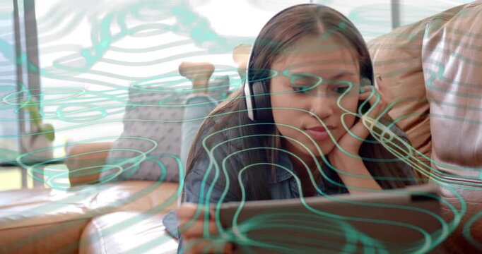 Teenage girl lying on sofa scrolling tablet adjusting headphones reacting to tech graphic overlay