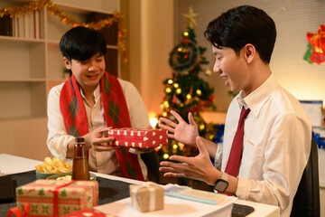 Two coworkers exchange gifts during a nighttime office Christmas party, surrounded by festive lights and decorations