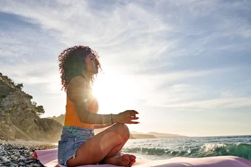 Fotobehang Lotusbloem Young woman with curly hair meditating in lotus pose on a yoga mat by the ocean, finding inner peace and balance  © Javier