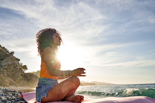 Young woman with curly hair meditating in lotus pose on a yoga mat by the ocean, finding inner peace and balance