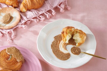 A freshly baked croissant is split open on a white plate, complemented by a golden spoon and a swirl of almond butter. Other pastries rest nearby on a light pink fabric