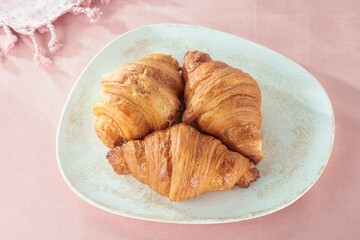 Three golden brown croissants rest on a speckled plate, set against a pastel pink background with a textured cloth, inviting thoughts of a Parisian morning