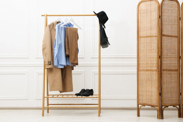 Different women's clothes hanging on rack and folding screen near white wall indoors