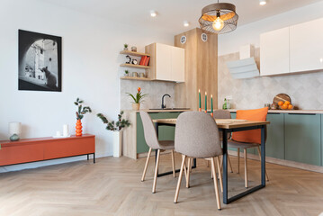 Modern kitchen and wooden dining table with chairs. Green furniture, beige tiles on the wall and orange decoration. New home with elegant design and picture on the wall. Real estate.	
