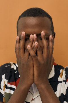 Young Black man covering face with both hands, standing against plain background, eyes closed, short hair visible, wearing patterned shirt, expressing emotion or stress
