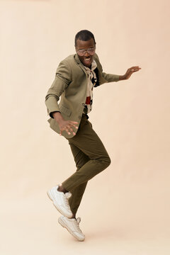 Young Black man jumping with energetic pose against plain background, wearing glasses and smiling, body angled sideways, arms extended, expressing dynamic movement