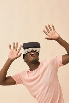 Black man wearing virtual reality headset raising hands and looking upward experiencing immersive digital simulation against plain background