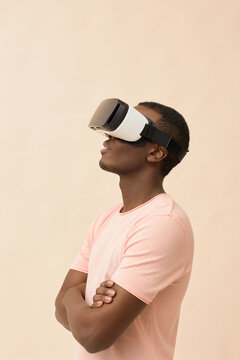 African American man standing with arms crossed wearing virtual reality headset, looking upward and engaging with immersive digital experience against plain background