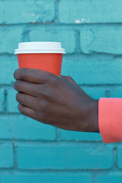 Close-up of Black person's hand in coral sleeve holding orange takeaway cup with white lid against vibrant blue brick wall creating bold urban contrast
