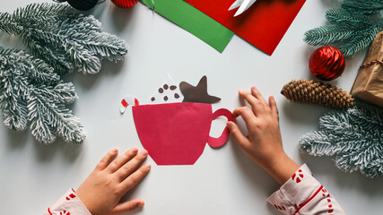 Child's hands crafting a paper cup with Christmas decorations on a white surface