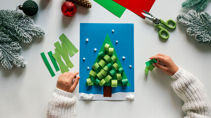 Child making a Christmas tree card with paper and decorations on a white table