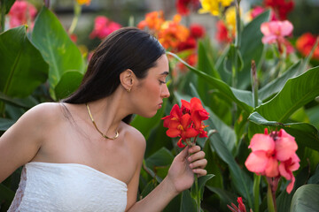 Fototapeta premium Young woman, beautiful, brunette, blue eyes, Hispanic, wearing a white top, surrounded by plants, smelling the scent of beautiful colorful flowers. Concept: model, beauty, fashion, trendy.
