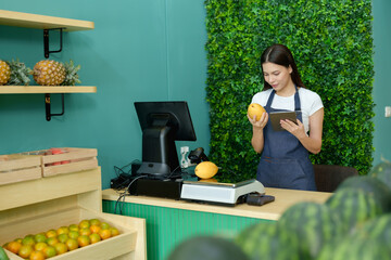 Caucasian female fruit shopkeeper preparing store for opening using tablet application to scan mango information while standing behind organized grocery counter filled with tropical fresh produce