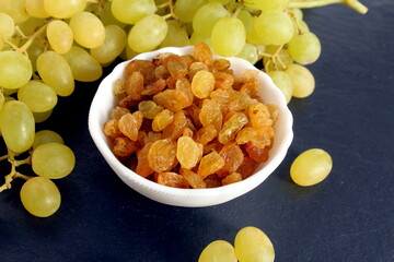 On a black background there is a white bowl with yellow raisins and fresh grapes.