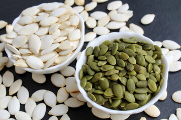 On a black background stand two bowls with peeled and unpeeled pumpkin seeds.	