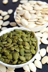 On a black background stand two bowls with peeled and unpeeled pumpkin seeds.	