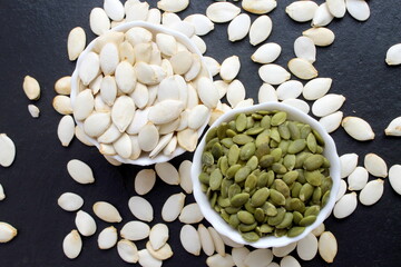 On a black background stand two bowls with peeled and unpeeled pumpkin seeds.