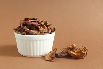 On a brown background lies a small pile of dried apple pieces in a bowl.	