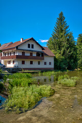 Rastoke, Croatia – July 21, 2025: A traditional wooden and stone mill house stands beside a rustic footbridge leading over the river in the village of Rastoke.
