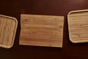 Overhead arrangement of three natural bamboo boards (one cutting board, two serving trays) set against a dark wood background, emphasizing wood grain and kitchen essentials.