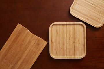 Abstract top-down view of bamboo cutting boards and square serving trays arranged on a rich dark brown wood background, emphasizing texture and natural materials.
