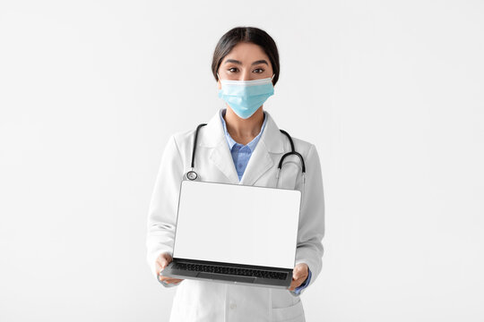 A millennial Hindu woman dressed in a coat is holding a laptop with an empty screen. She is isolated against a white background, promoting online consultations with healthcare professionals.