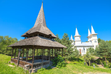 Traditional wooden gazebo with a tall shingled roof in the foreground, historic Saint Parascheva Romanian Orthodox church and spire visible behind in Poienile Izei, Maramures in Romania
