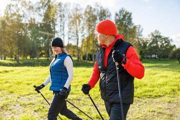 Fototapeta premium A man and a woman dressed in sporty autumn gear walk with Nordic poles across a grassy park on a bright, sunny day, enjoying fitness in nature